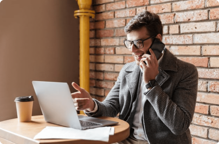 Happy looking man on his phone while looking at his laptop and sitting in a cafe